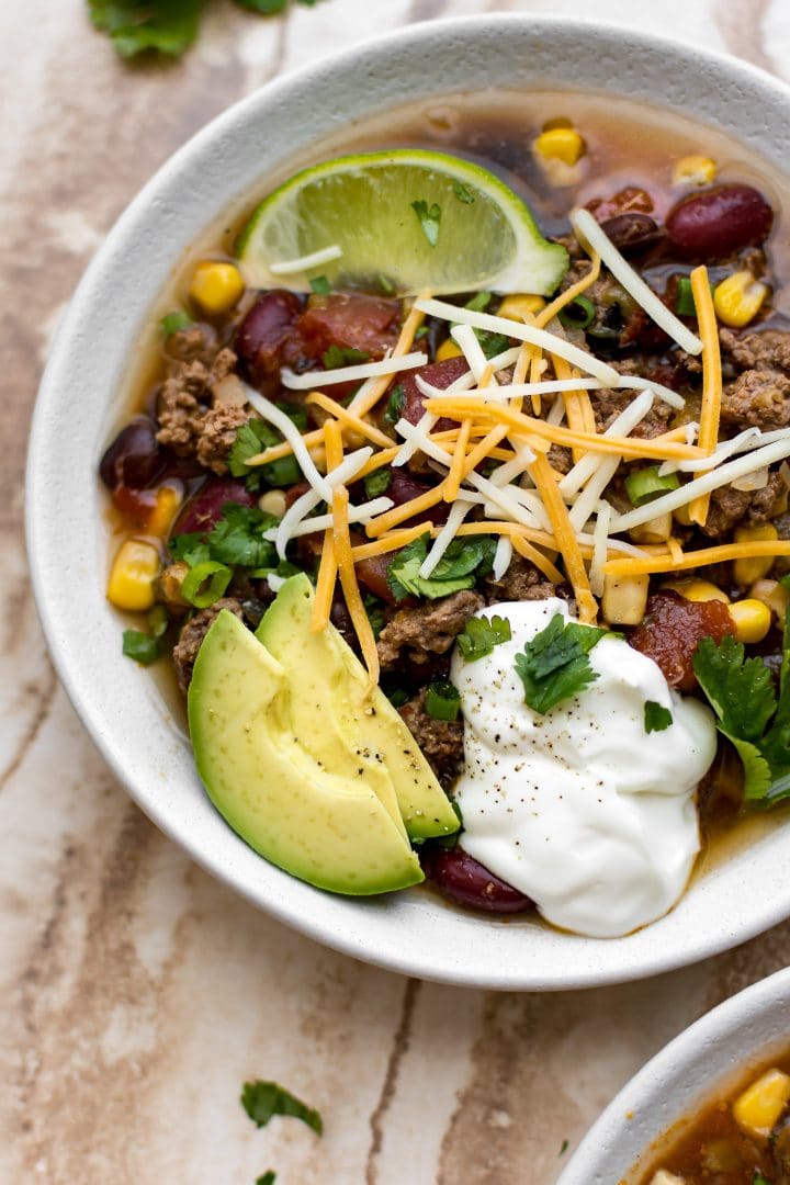 close-up of a white bowl with slow cooker beef taco soup recipe topped with lime, avocado, and sour cream