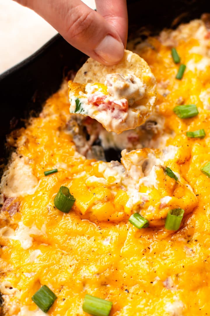 cheesy sausage dip close-up with Natasha scooping some up with a round tortilla chip