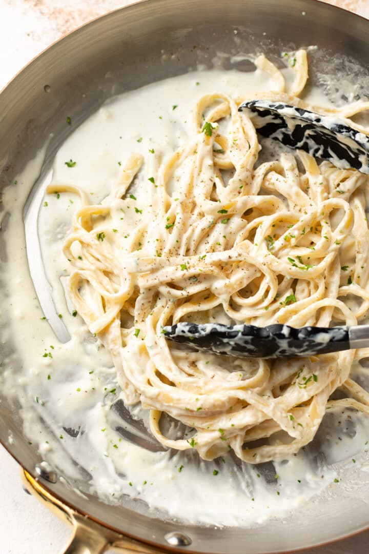 a skillet with fettuccine alfredo and serving tongs