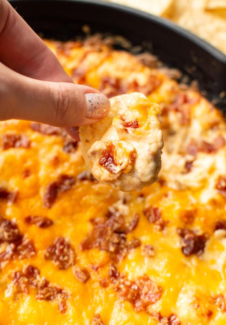 holding a chip scooping cheddar bacon dip from skillet