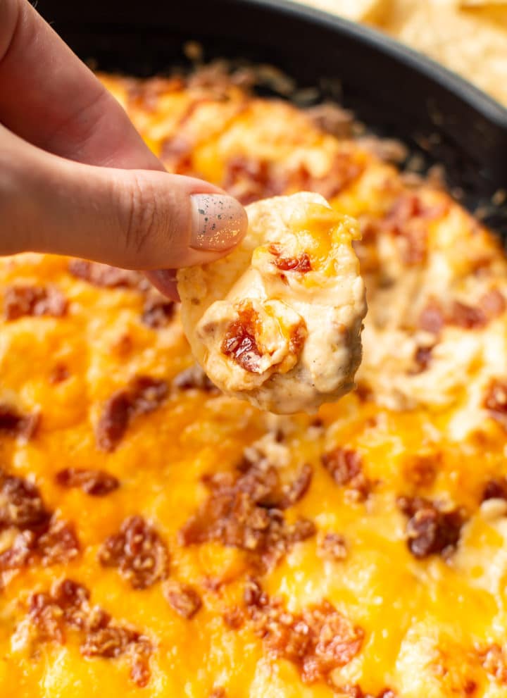 holding a chip scooping cheddar bacon dip from skillet