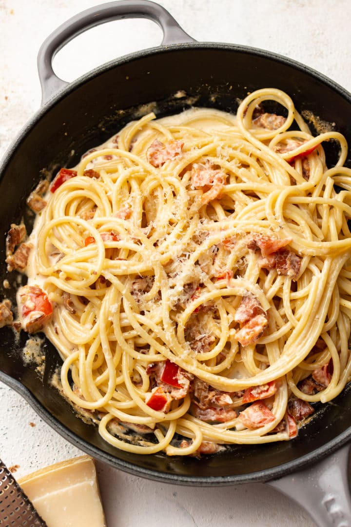 bacon and tomato pasta in a skillet with a wedge of parmesan and a cheese grater beside it