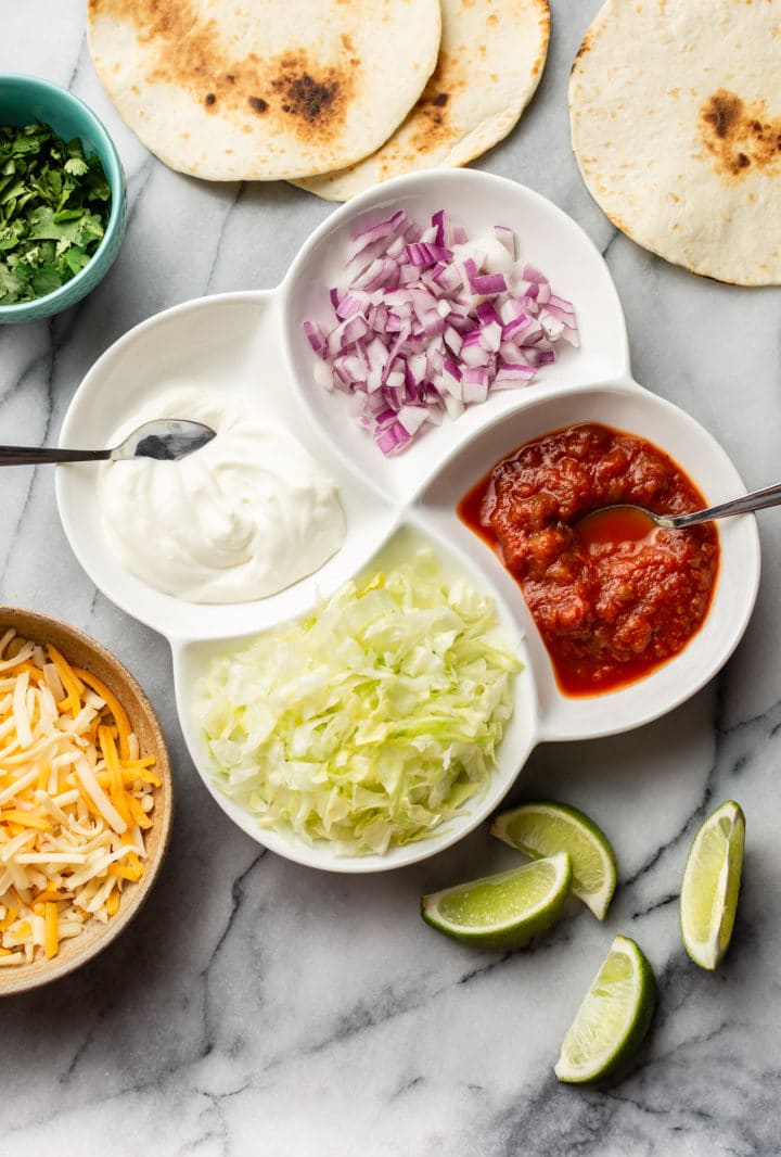 toppings for ground beef tacos on a marble background (salsa, sour cream, lime wedges, red onions, cilantro, cheese blend, iceberg lettuce)