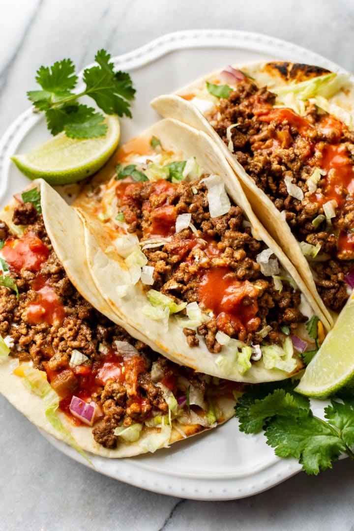 close-up of three ground beef tacos on a white plate with extra lime wedges and cilantro