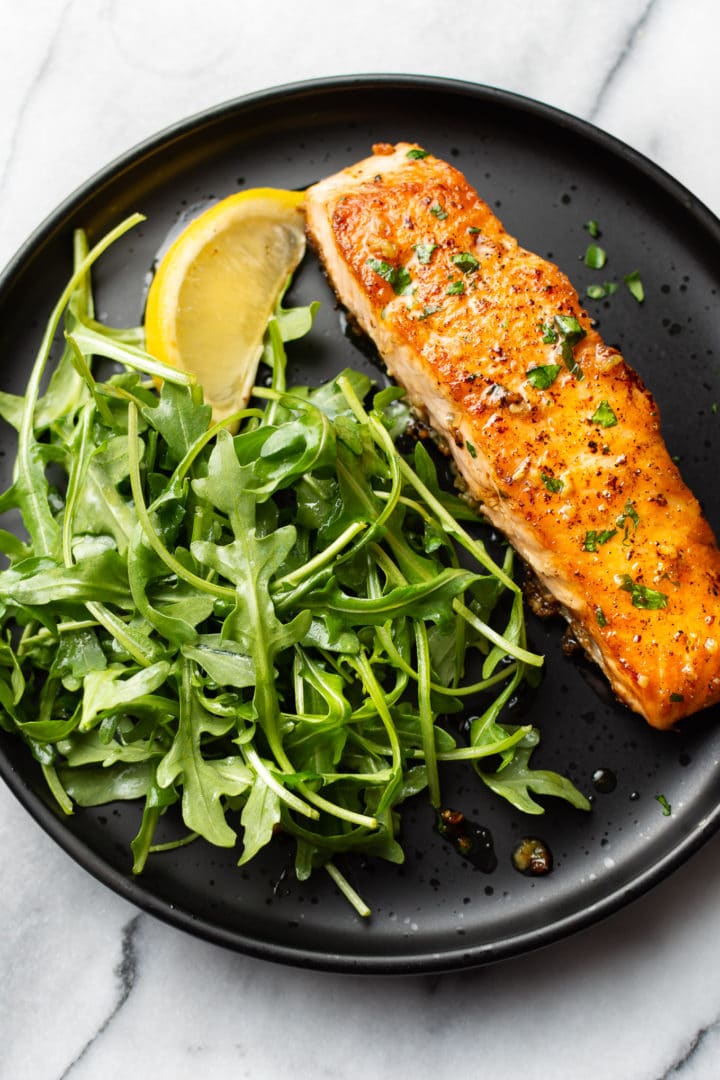 garlic butter salmon on a black plate with arugula salad and a lemon wedge