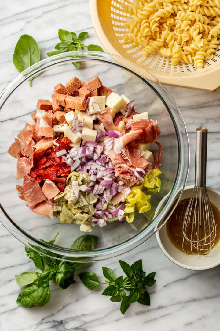 Italian antipasto salad in a glass prep bowl pictured with homemade dressing, fresh basil, and cooked pasta