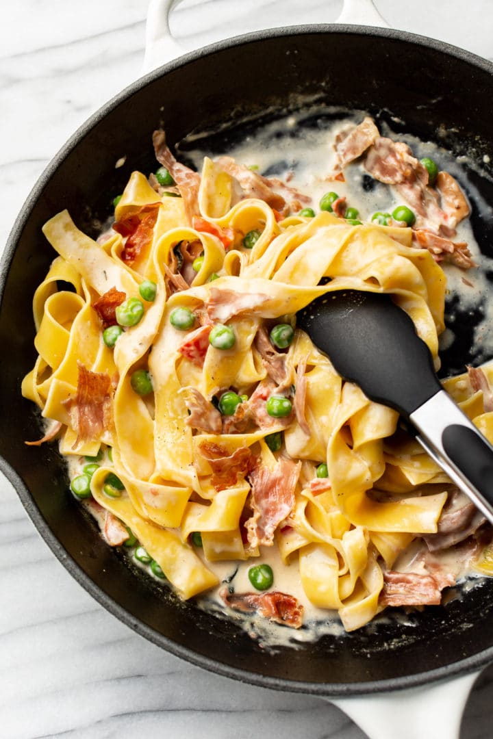 pasta with prosciutto and peas in a skillet being tossed with tongs