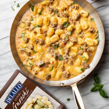 spicy Italian sausage gnocchi skillet on a marble background surrounded by fresh basil, a package of DeLallo potato gnocchi, and a jar of DeLallo chopped calabrian chili peppers