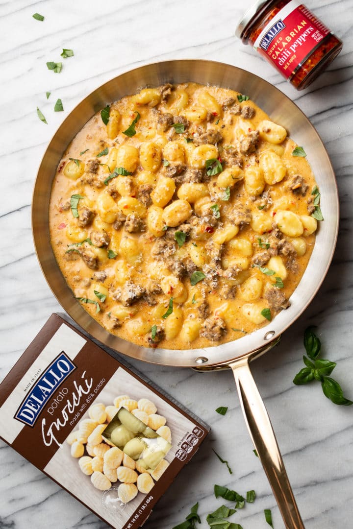 spicy Italian sausage gnocchi skillet on a marble background surrounded by fresh basil, a package of DeLallo potato gnocchi, and a jar of DeLallo chopped calabrian chili peppers