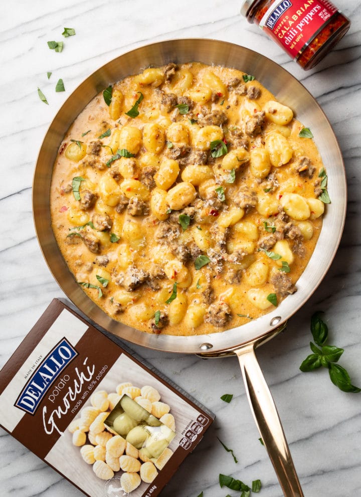 spicy Italian sausage gnocchi skillet on a marble background surrounded by fresh basil, a package of DeLallo potato gnocchi, and a jar of DeLallo chopped calabrian chili peppers