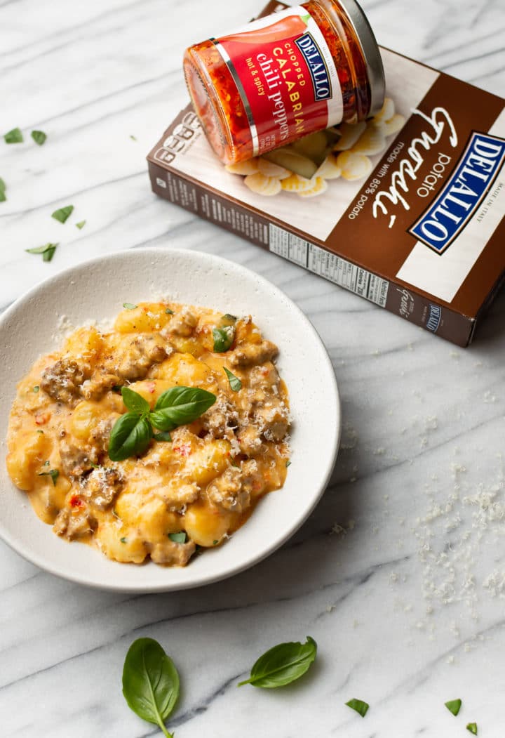 spicy Italian sausage gnocchi in a white bowl surrounded by fresh basil, a package of DeLallo potato gnocchi, and a jar of DeLallo calabrian chili peppers