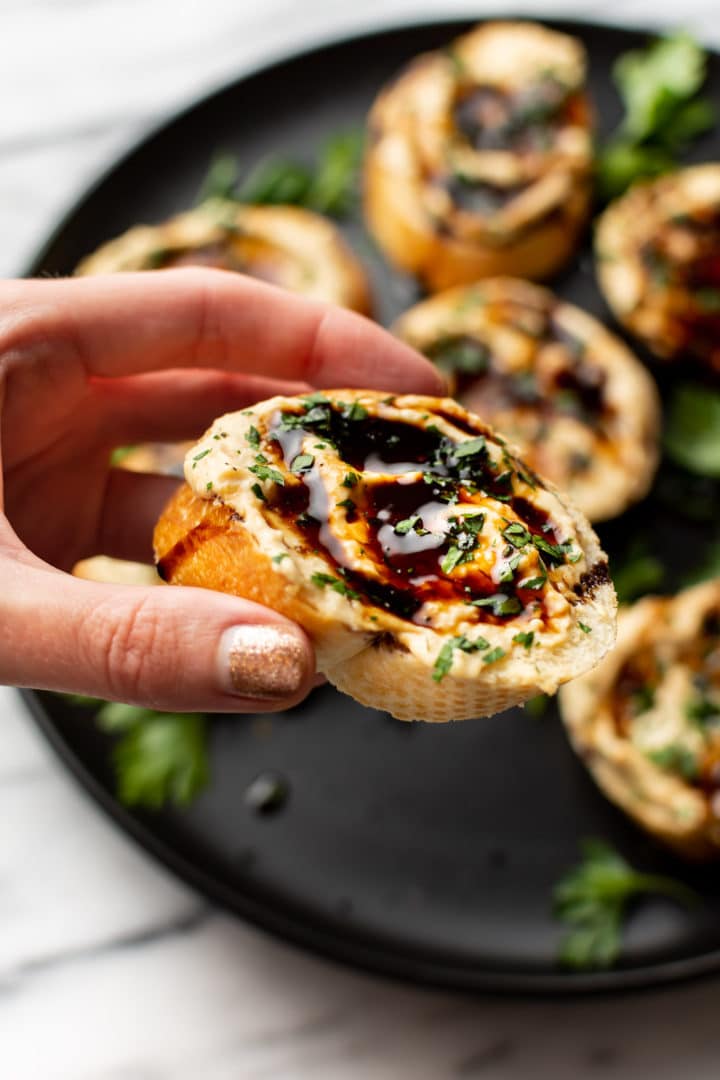 a feminine hand lifting up a piece of hummus crostini with the platter in the background