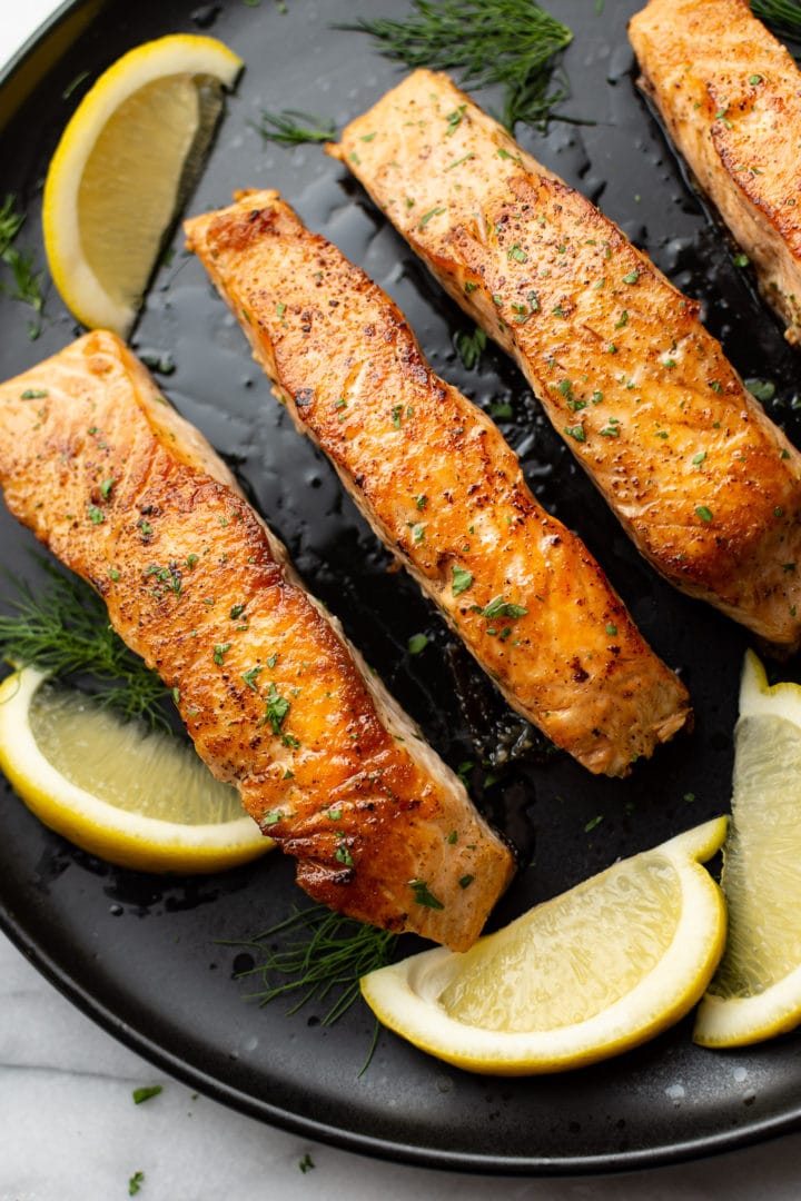 pan seared salmon on a black plate surrounded by lemon wedges