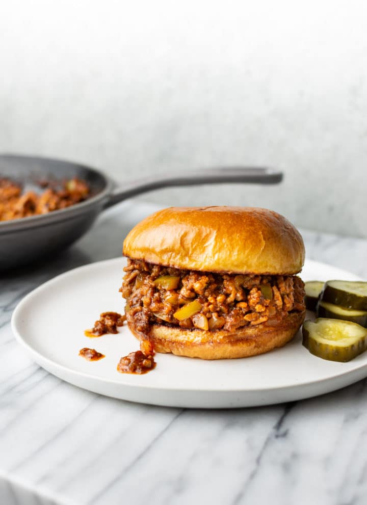 Sloppy Joe sandwich on a white plate with pickles and the skillet in the background