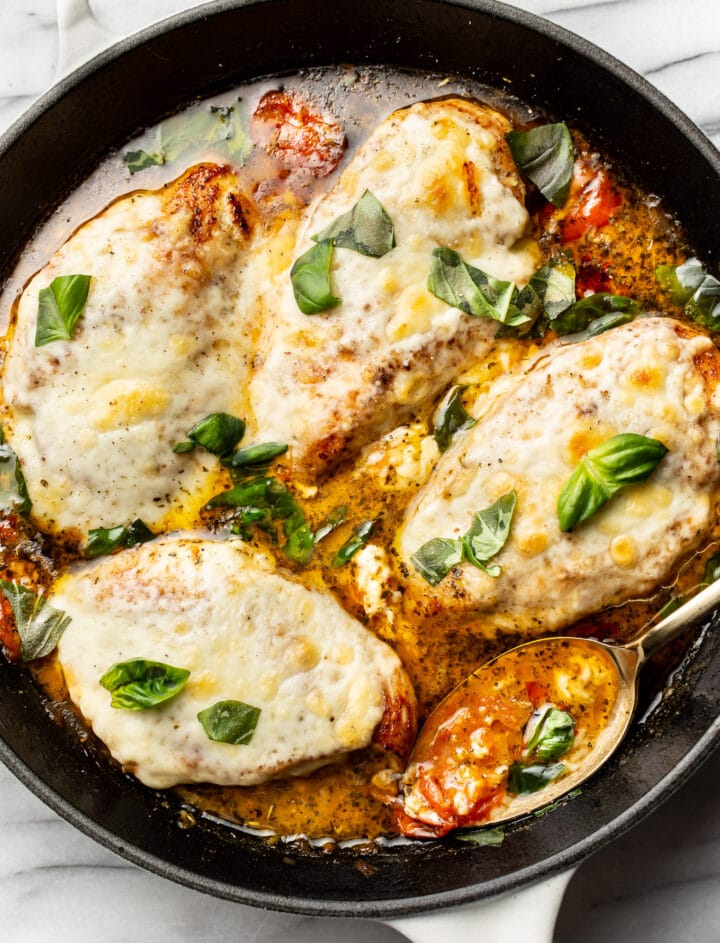 a cast iron skillet with four pieces of pesto chicken in sauce garnished with basil and mozzarella