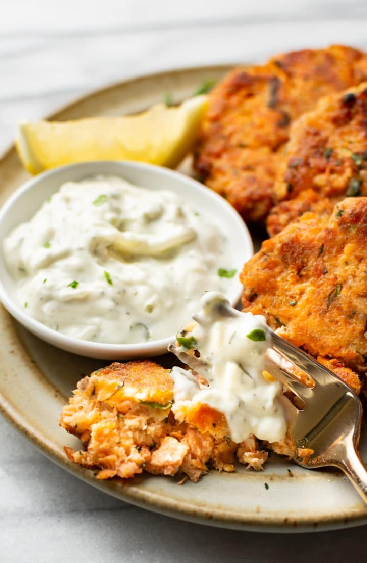 close-up of a plate with salmon cakes, dip, and a lemon wedge next to a forkful of salmon patty