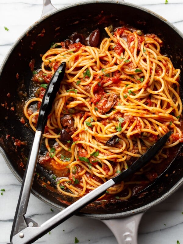 a skillet with spaghetti in puttanesca sauce with serving tongs