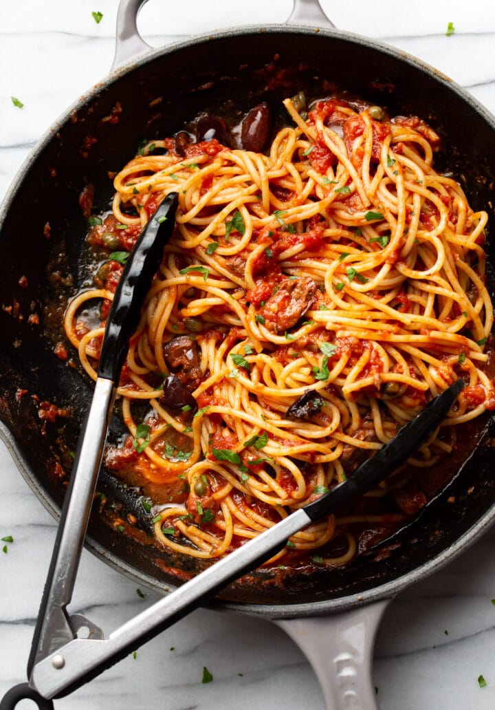 a skillet with spaghetti in puttanesca sauce with serving tongs