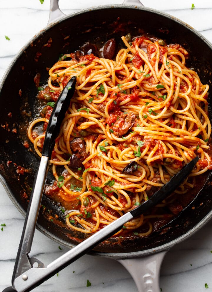 a skillet with spaghetti in puttanesca sauce with serving tongs