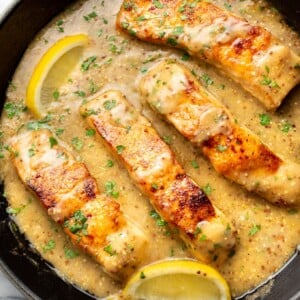 a cast iron skillet with four pieces of salmon in sauce with lemon wedges