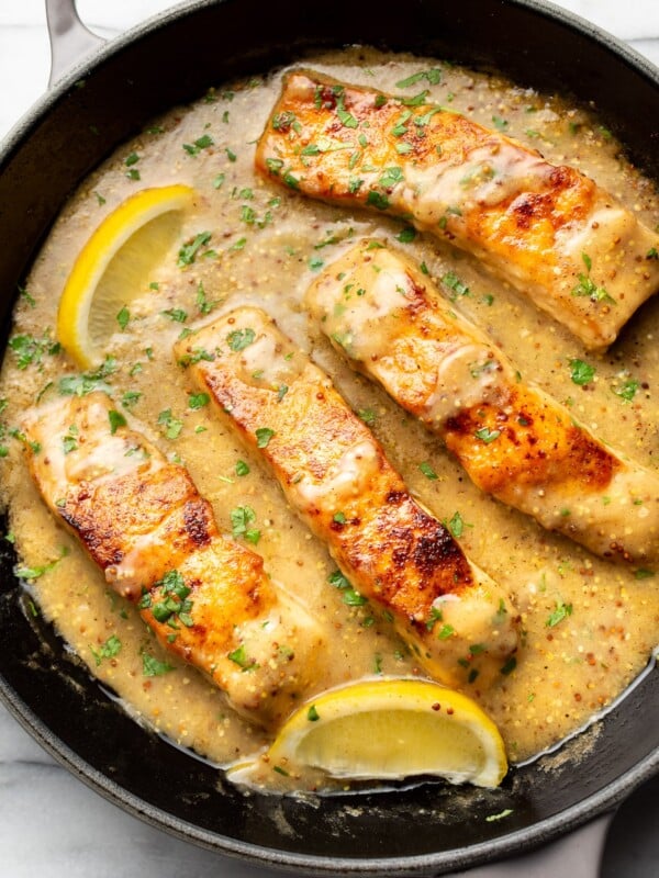 a cast iron skillet with four pieces of salmon in sauce with lemon wedges