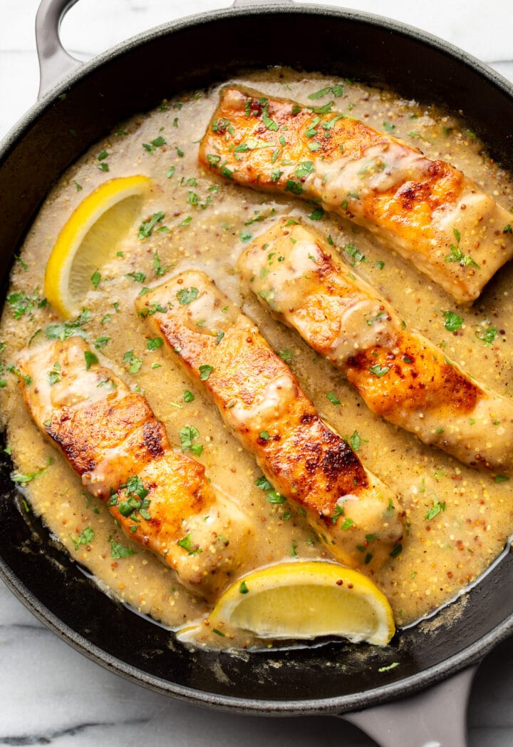 a cast iron skillet with four pieces of salmon in sauce with lemon wedges