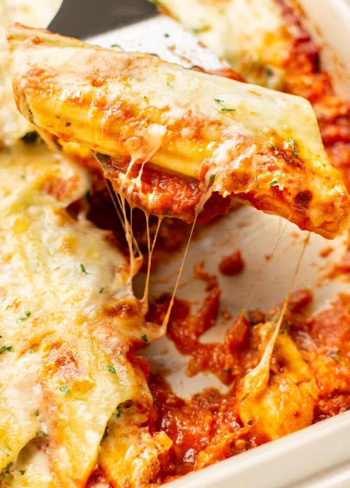 closeup of a metal spatula with cheesy manicotti above a baking dish