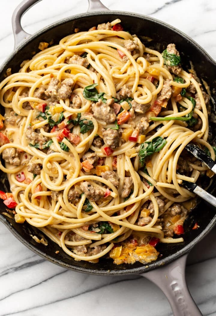 a skillet with creamy sausage and peppers pasta and serving tongs