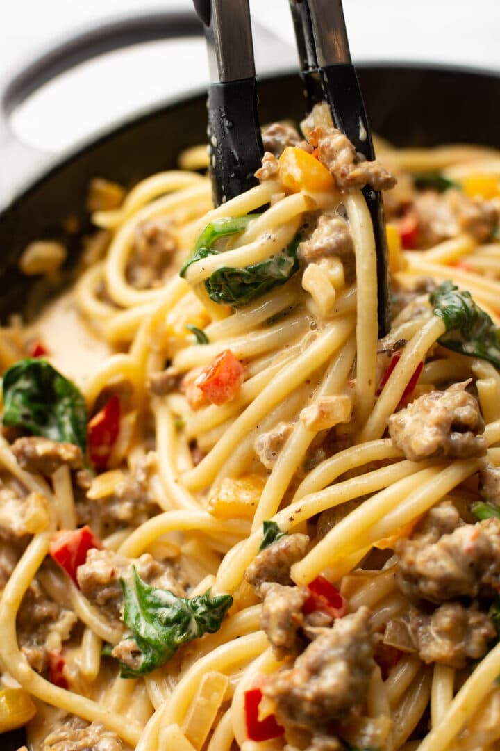 closeup of serving tongs with bucatini with sausage and peppers