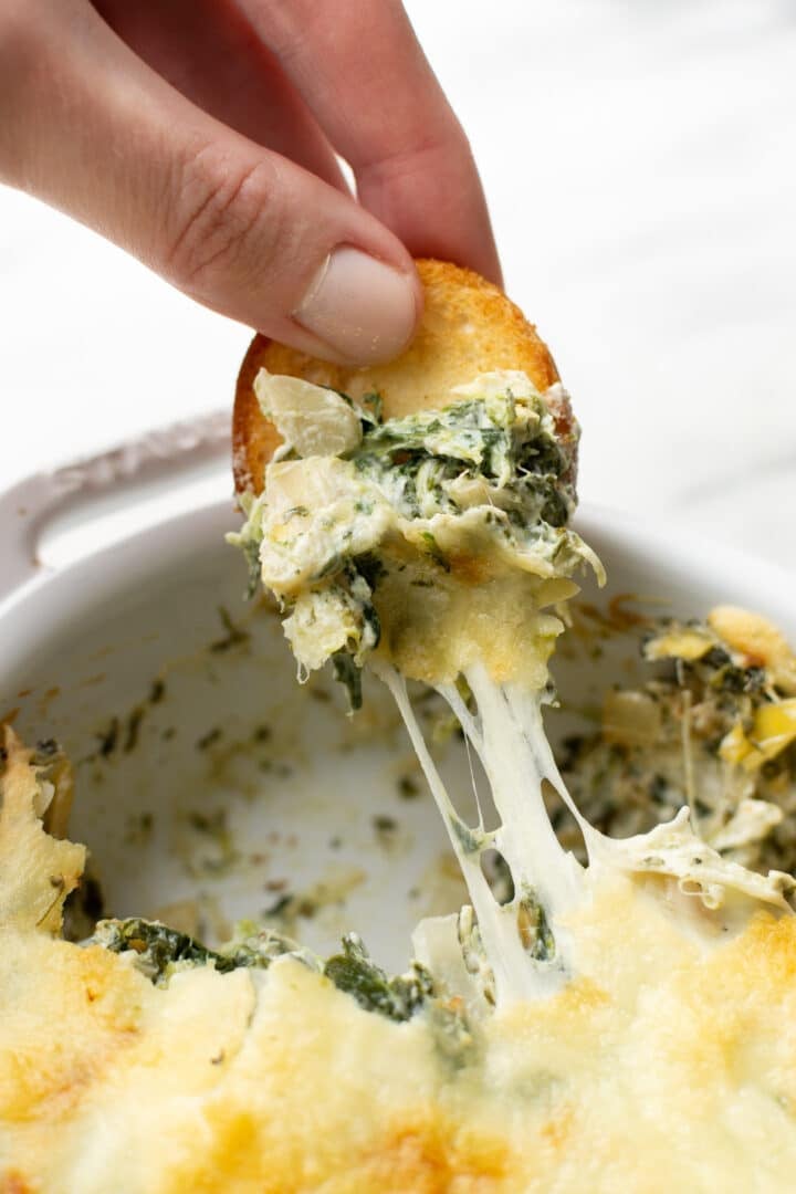 closeup of a hand dipping bread in spinach artichoke dip