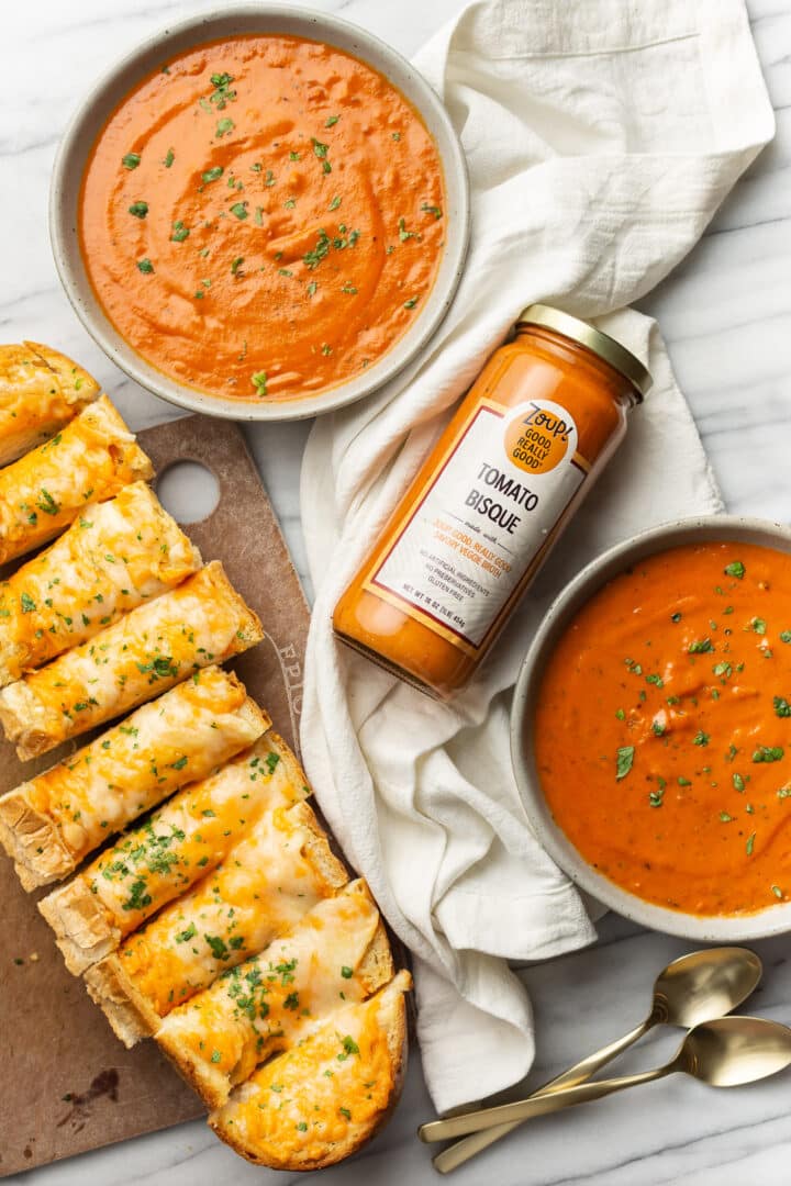 two bowls of tomato bisque next to half a loaf of cheesy bread and a jar of zoup