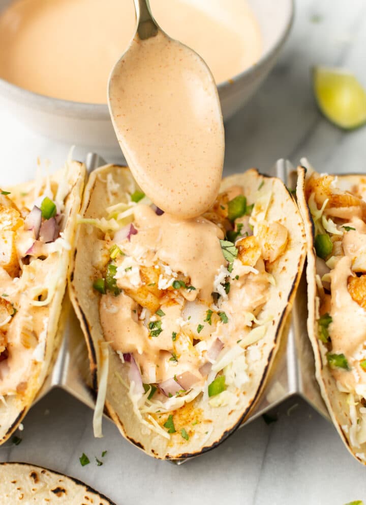 spooning sauce over three fish tacos