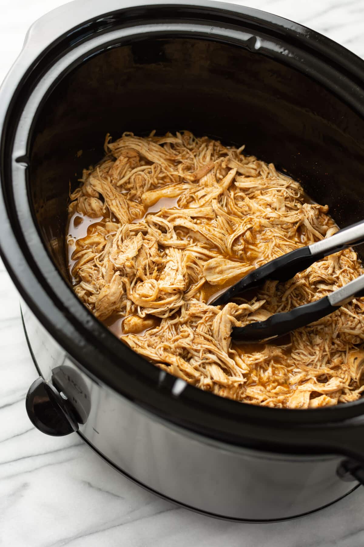 a slow cooker with shredded chicken and kitchen tongs