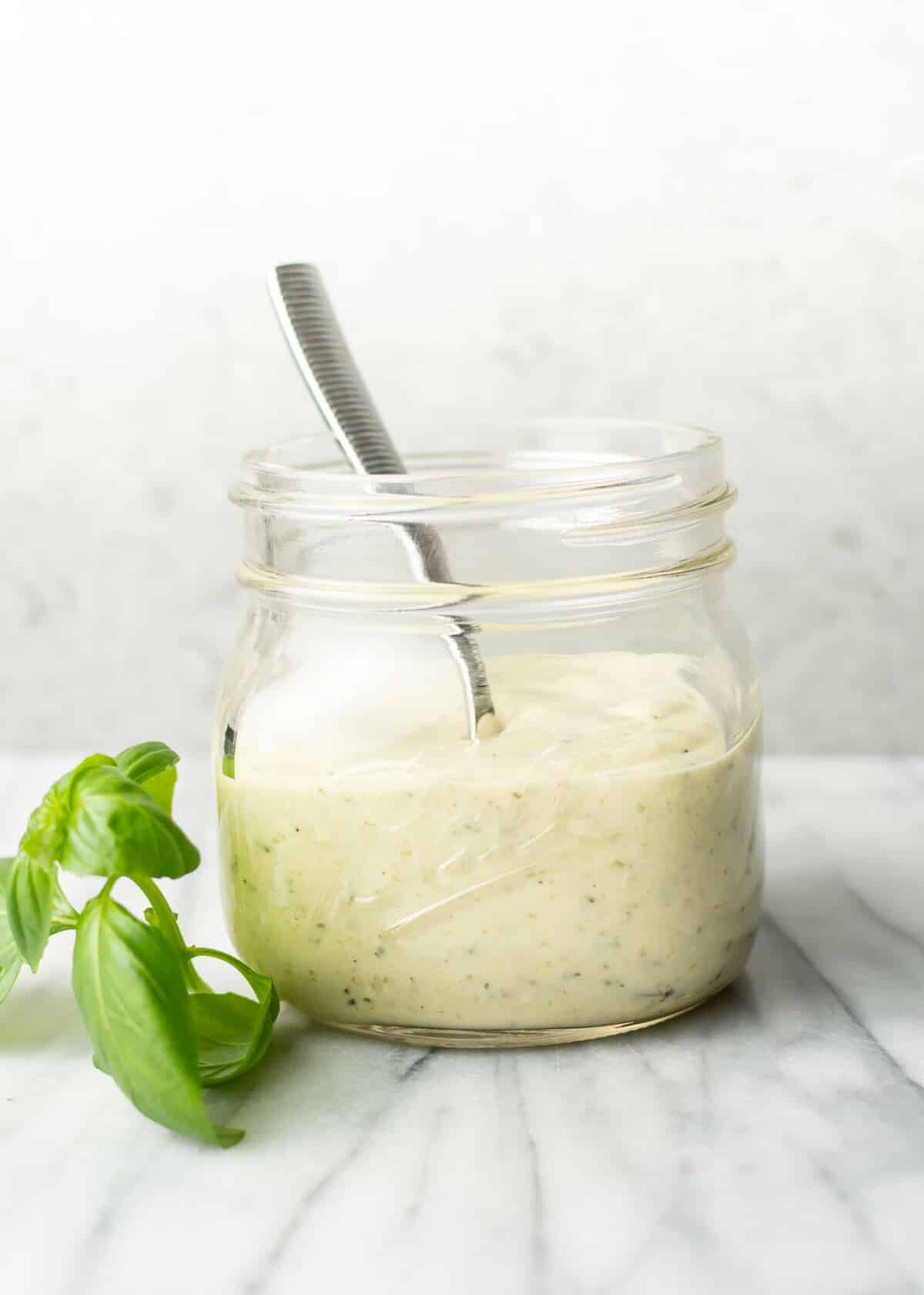 a glass jar with creamy pesto dressing, a spoon, and a sprig of basil