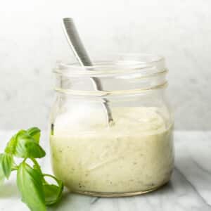 a glass jar with creamy pesto dressing, a spoon, and a sprig of basil