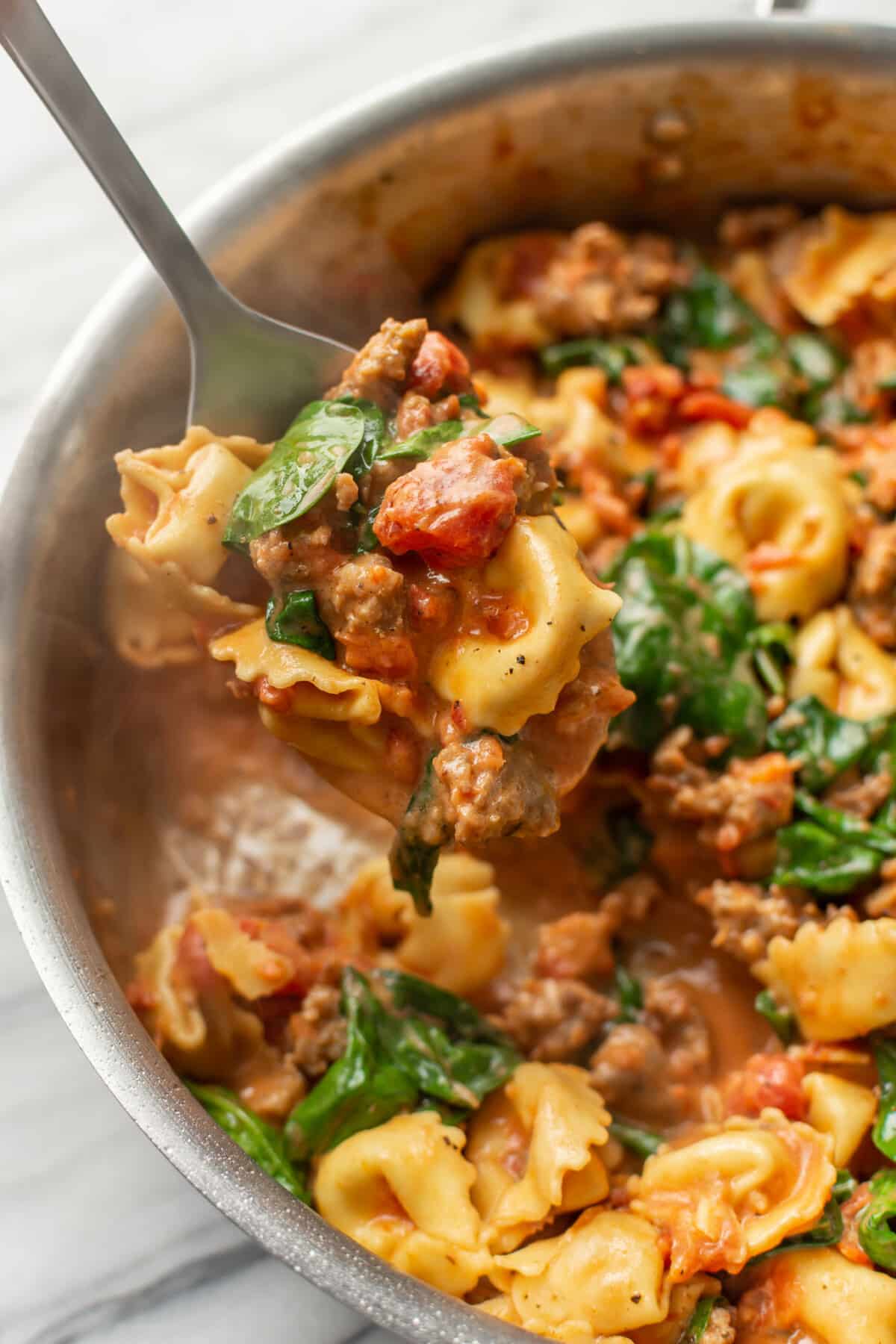 a skillet with cheese tortellini and italian sausage and a serving spoon