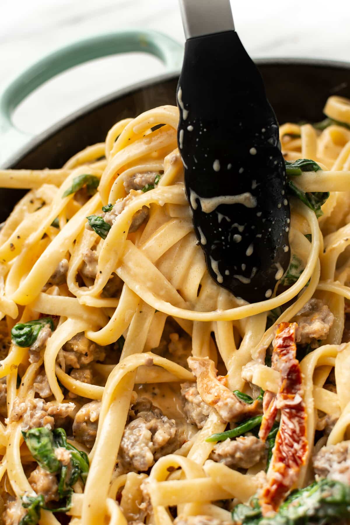 closeup of serving tongs with creamy tuscan sausage pasta in a skillet