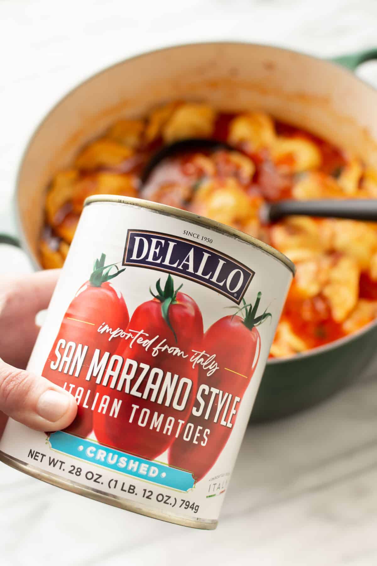 a pot of ravioli soup with a ladle and a female hand holding up a can of crushed tomatoes