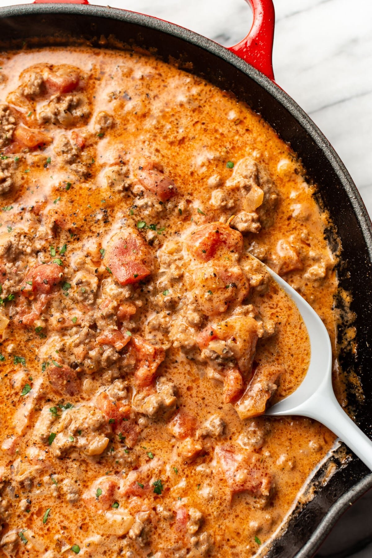 closeup of a skillet with creamy ground beef and tomato pasta sauce and a serving spoon