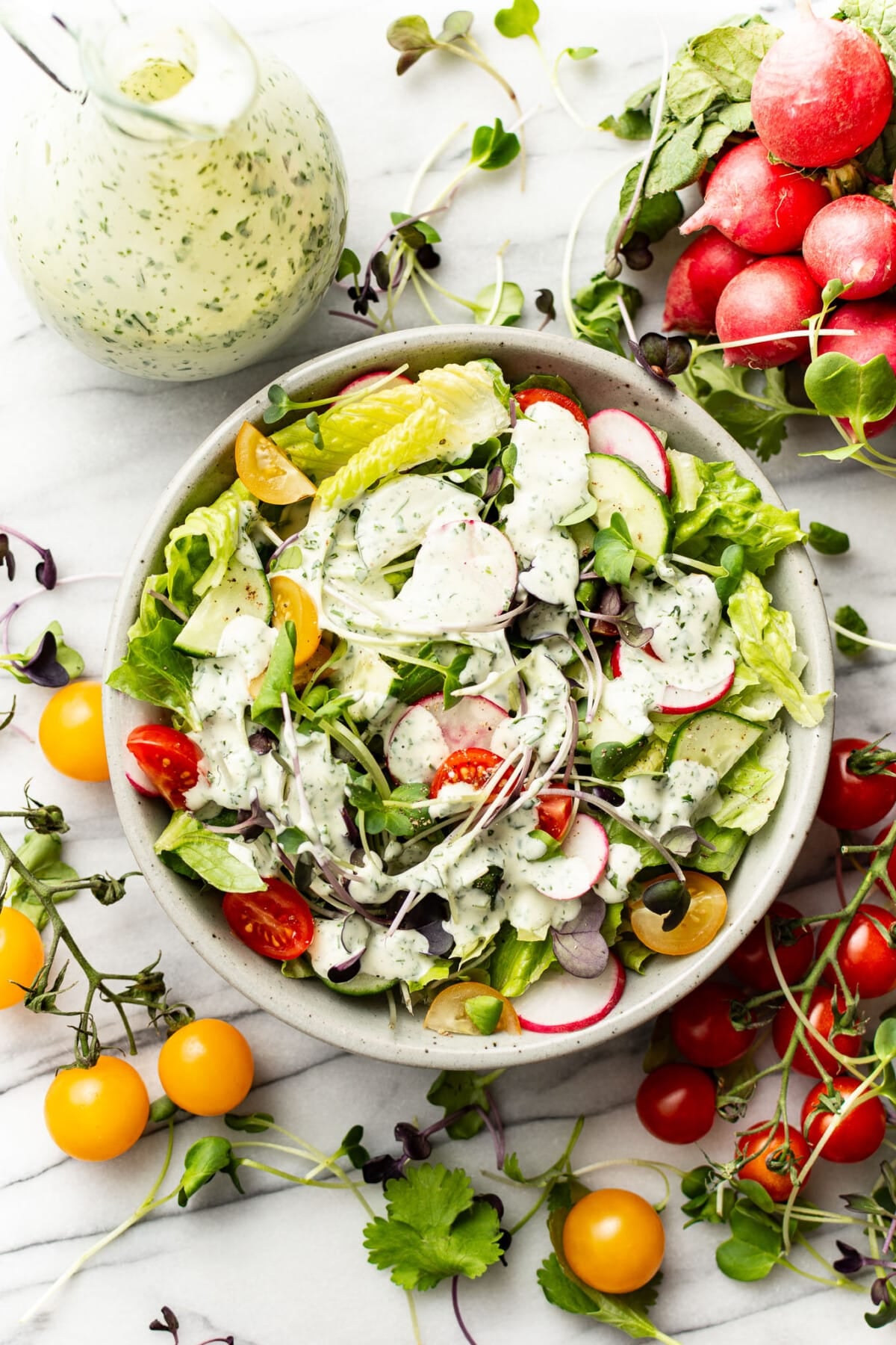 a bowl of salad next to a bottle with homemade green goddess dressing and tomatoes