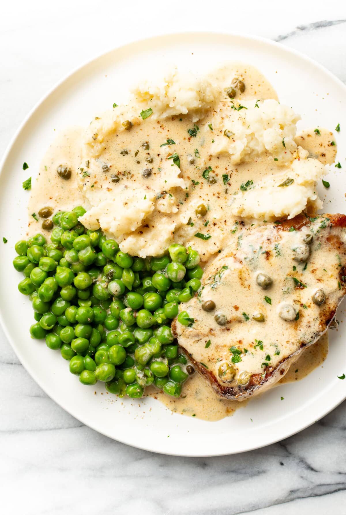 a plate with a pork chop in piccata sauce, mashed potatoes, and peas